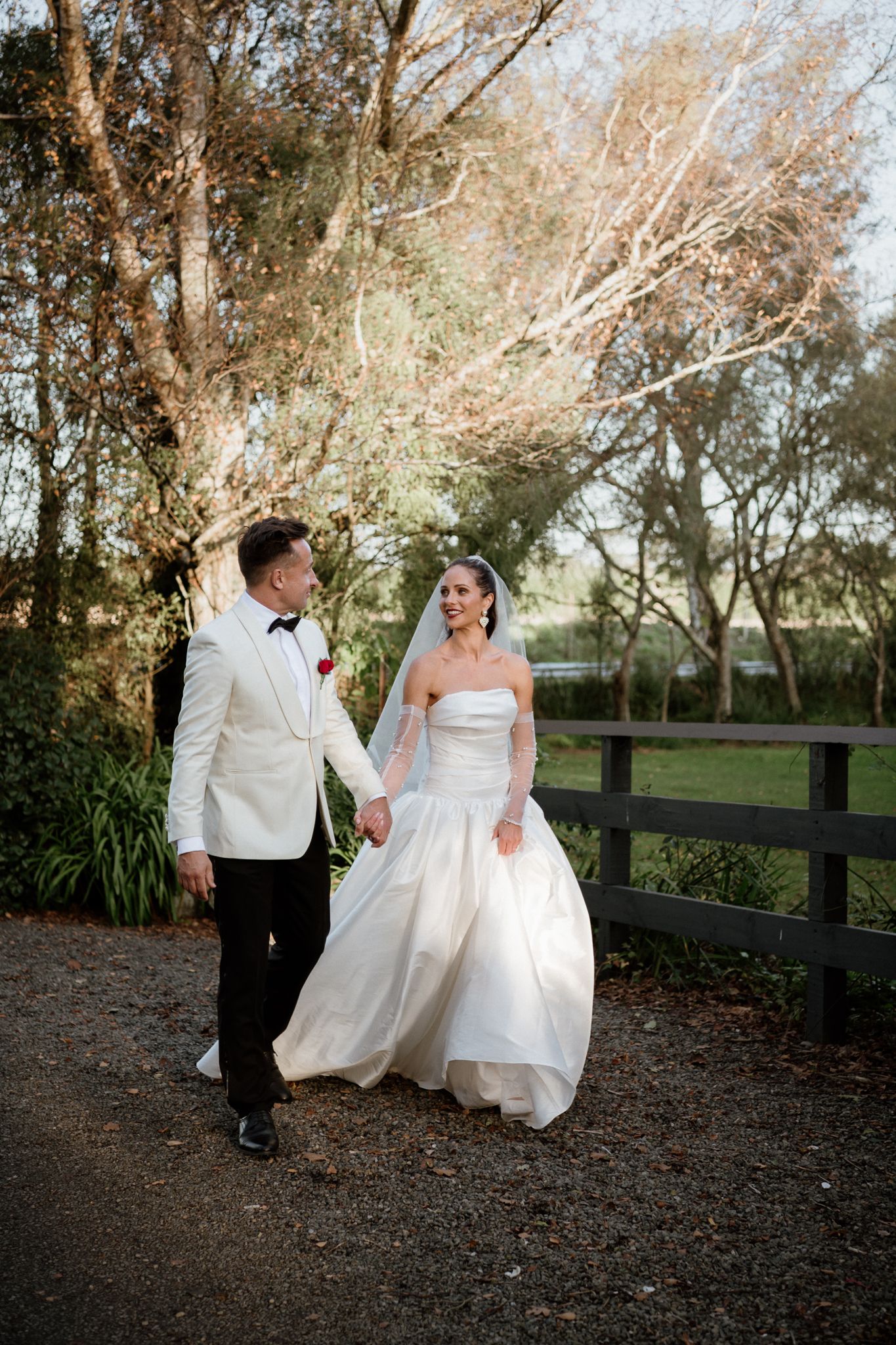 A bride in a wedding dress and groom in a white jacket walk down a tree-lined path at their Tarureka elopement by August and Vine