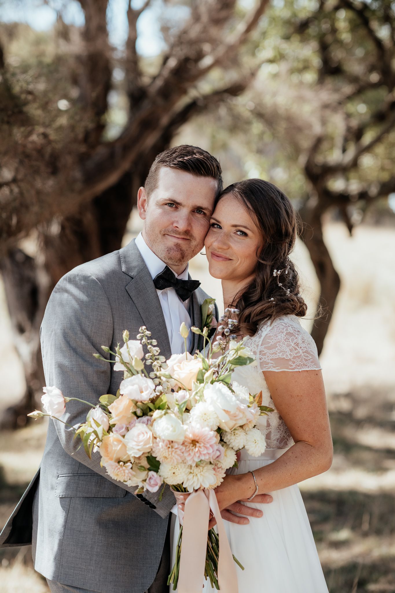 A joyful couple in wedding attire embrace outdoors holding a bouquet of blush and white flowers on their special day by August and Vine