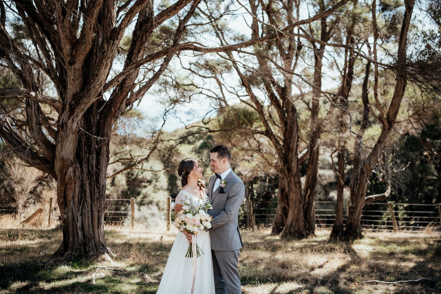 A couple in wedding attire embraces under rustic wooden trees during an outdoor ceremony at Whitimanuka by August and Vine