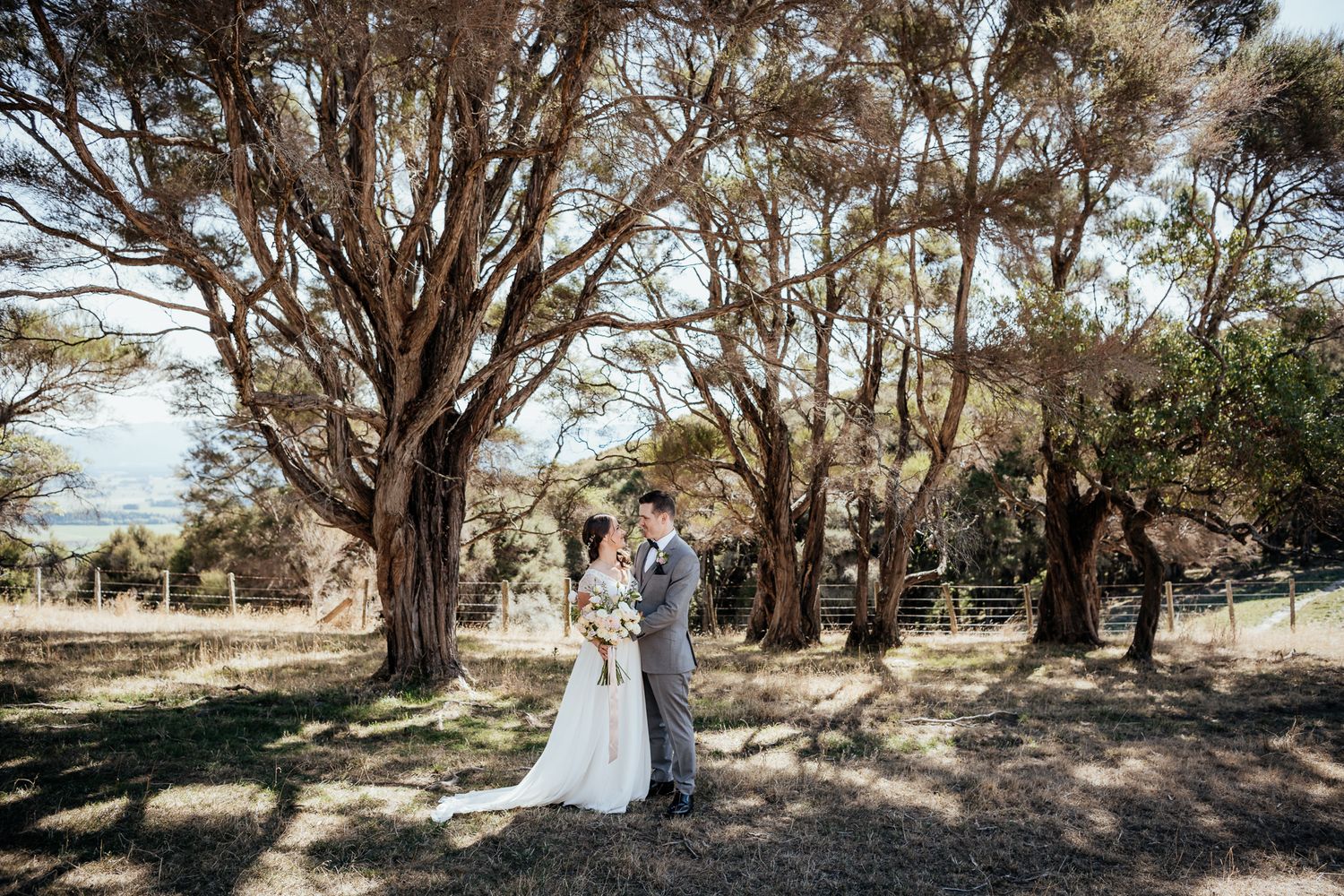 A romantic couple embraces under majestic Manuka on their outdoor wedding day at Whitimanuka by August and Vine