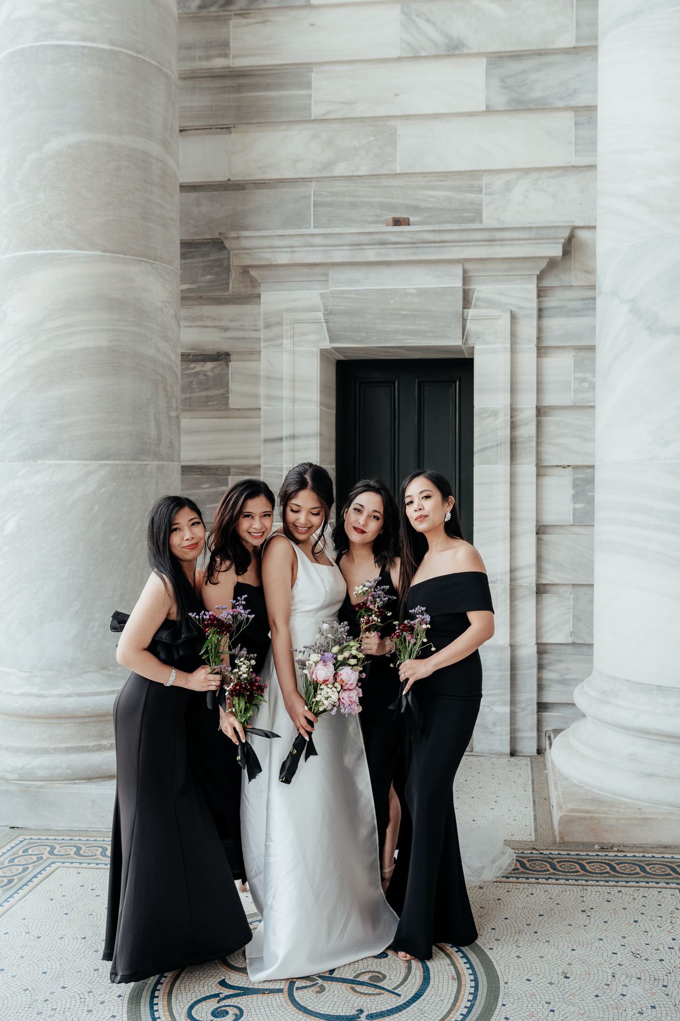 A bridal party in black dresses share a joyful moment with the bride between marble columns at NZ Parliament by August and Vine