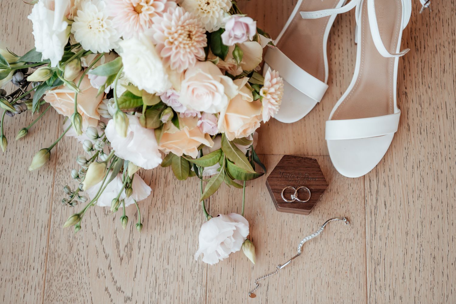 Wedding details with white bridal shoes, floral bouquet with roses and greenery, and golden wedding rings on wooden floor by August and Vine