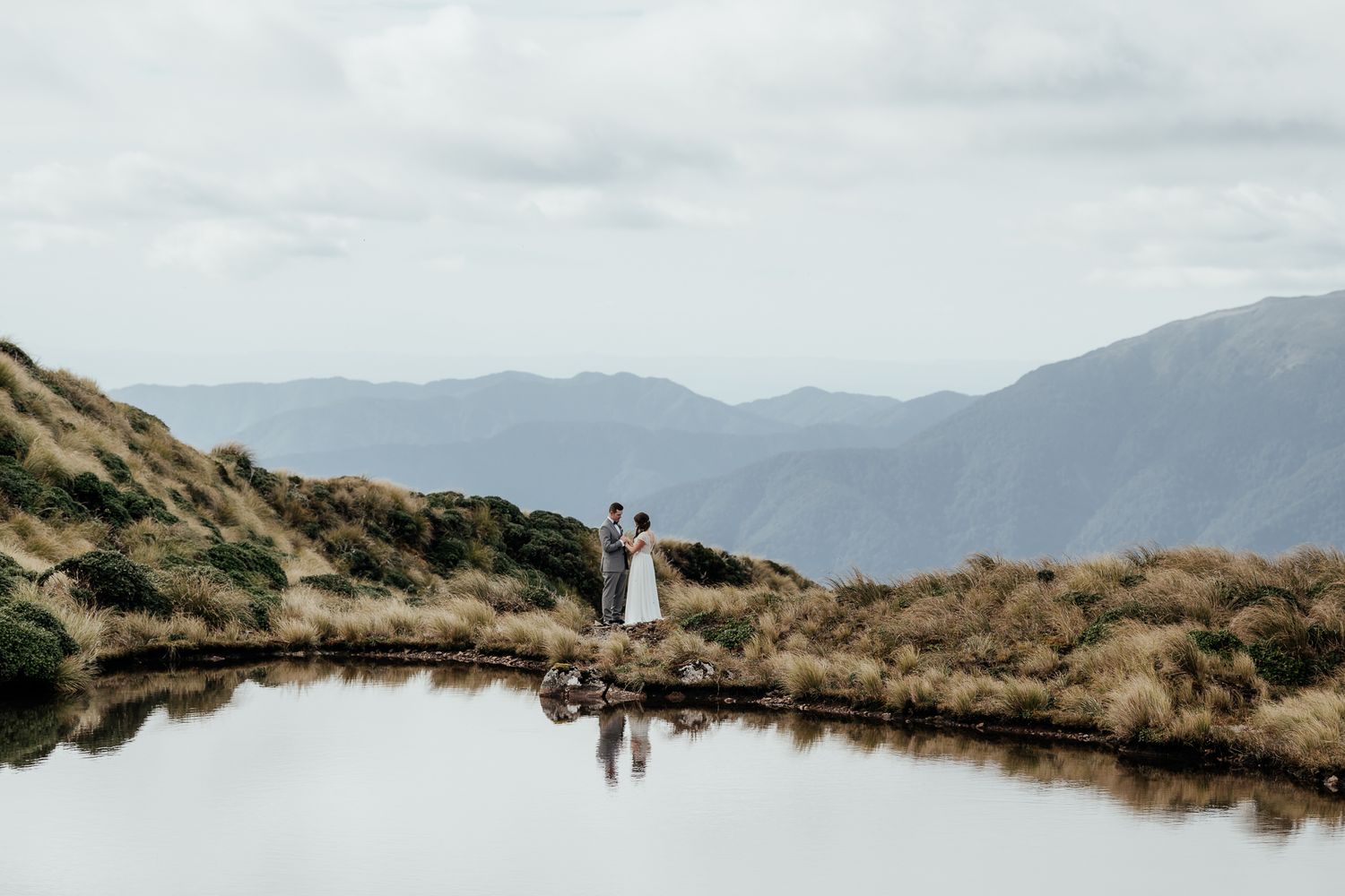 A couple stands near a serene mountain lake reflecting the cloudy sky and distant peaks in the landscape of the Tararuas by August and Vine