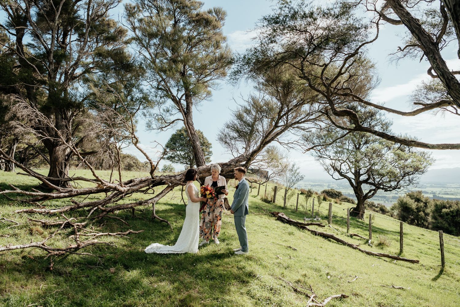 A wedding couple shares romantic moments under windswept trees on a scenic hillside during their elopement ceremony by August and Vine