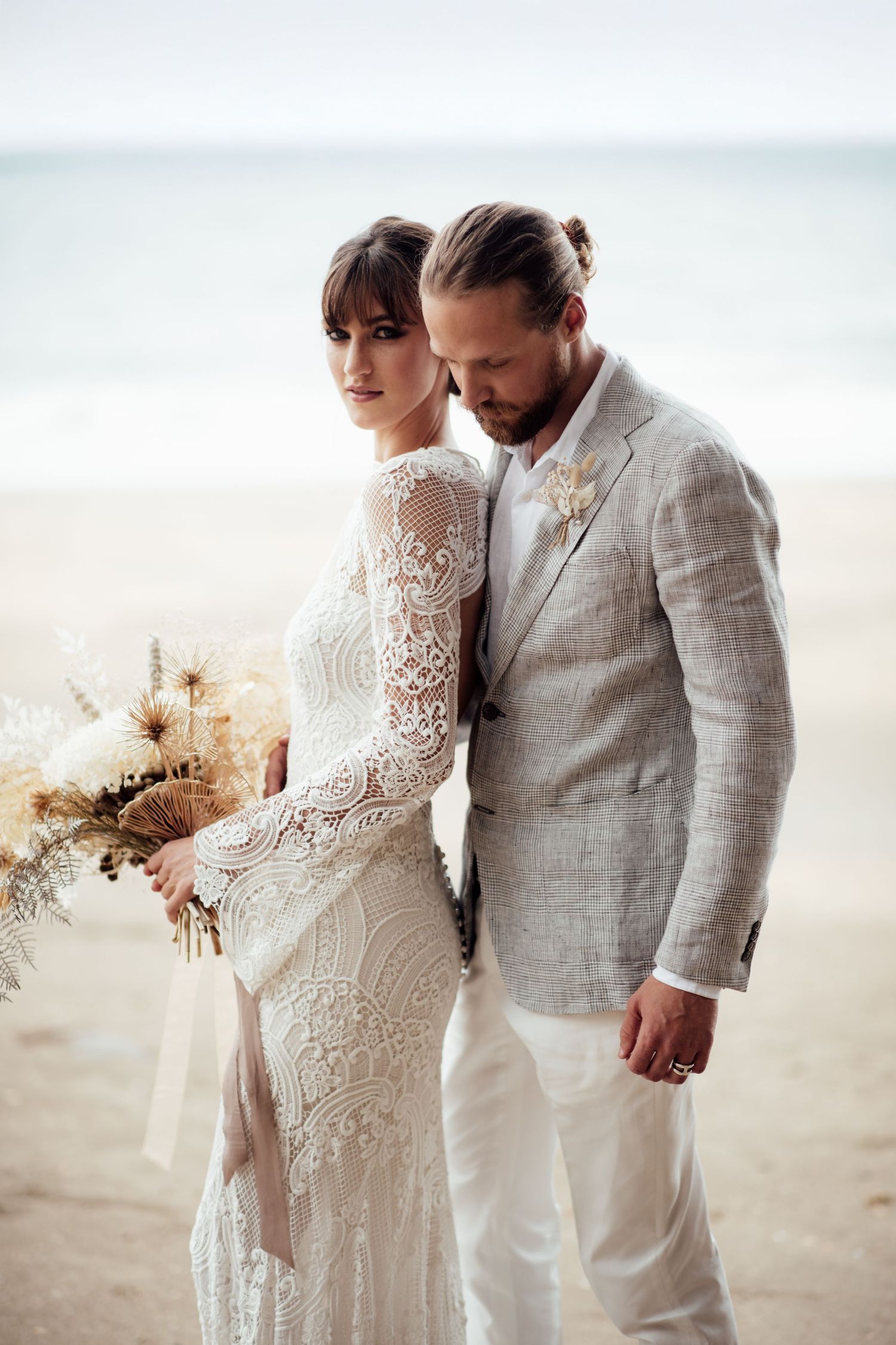 A romantic beach wedding scene with a couple in elegant white lace and linen attire standing close together by the ocean by August and Vine