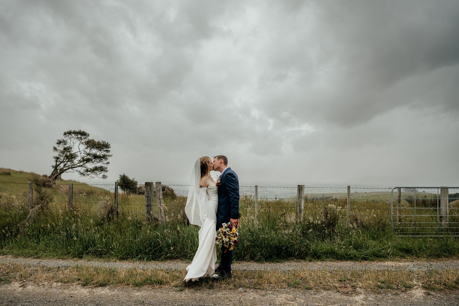 A couple in wedding attire embrace on a rural path under dramatic stormy clouds in the Wairarapa by August and Vine