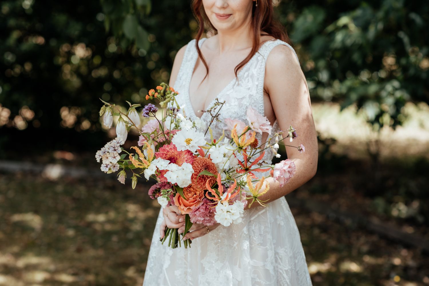 A bride with bouquet featuring vibrant coral and white flowers with cascading greenery against a natural outdoor backdrop by August and Vine