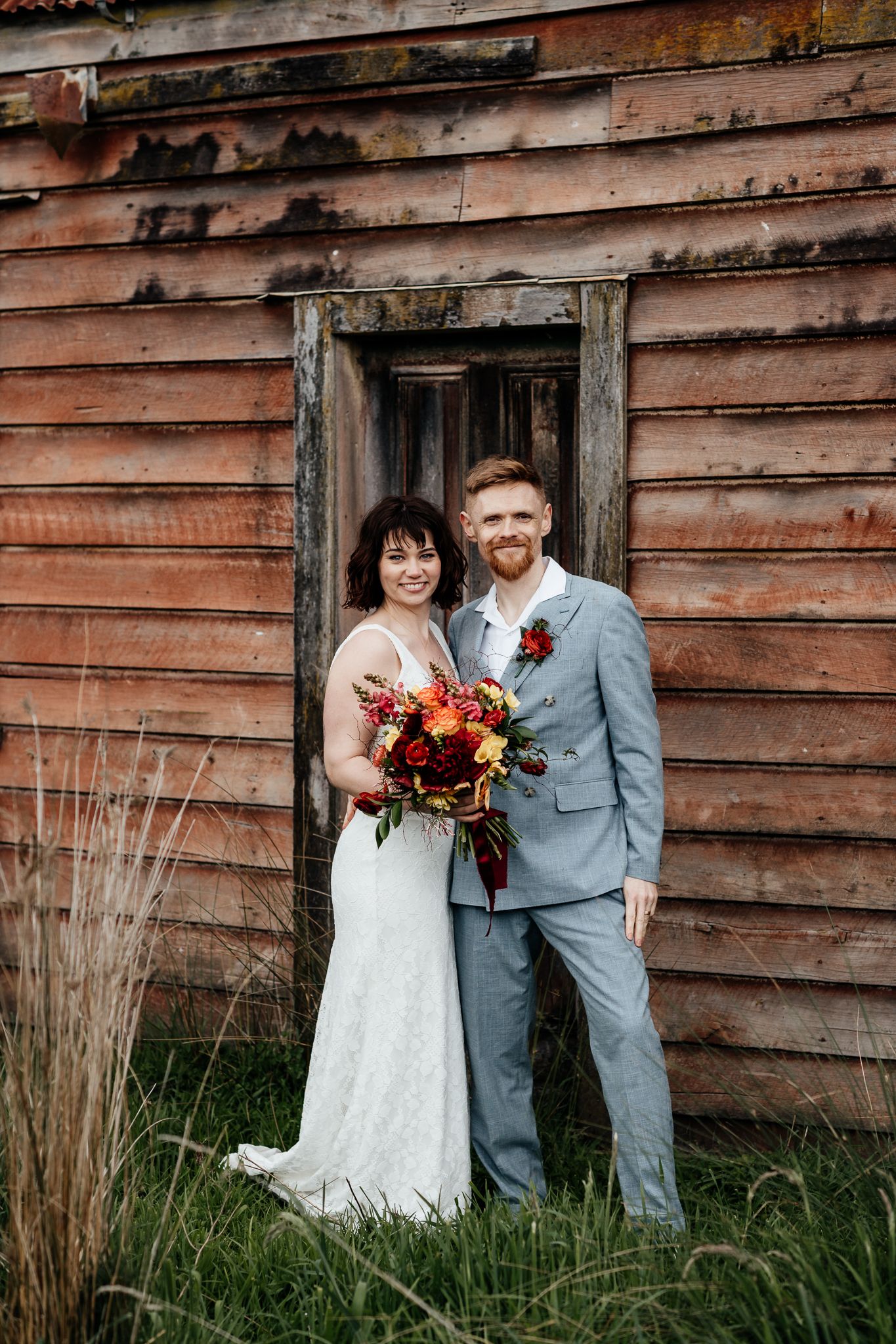 A couple in wedding attire stands against a rustic wooden Wairarapa barn surrounded by tall grass by August and Vine