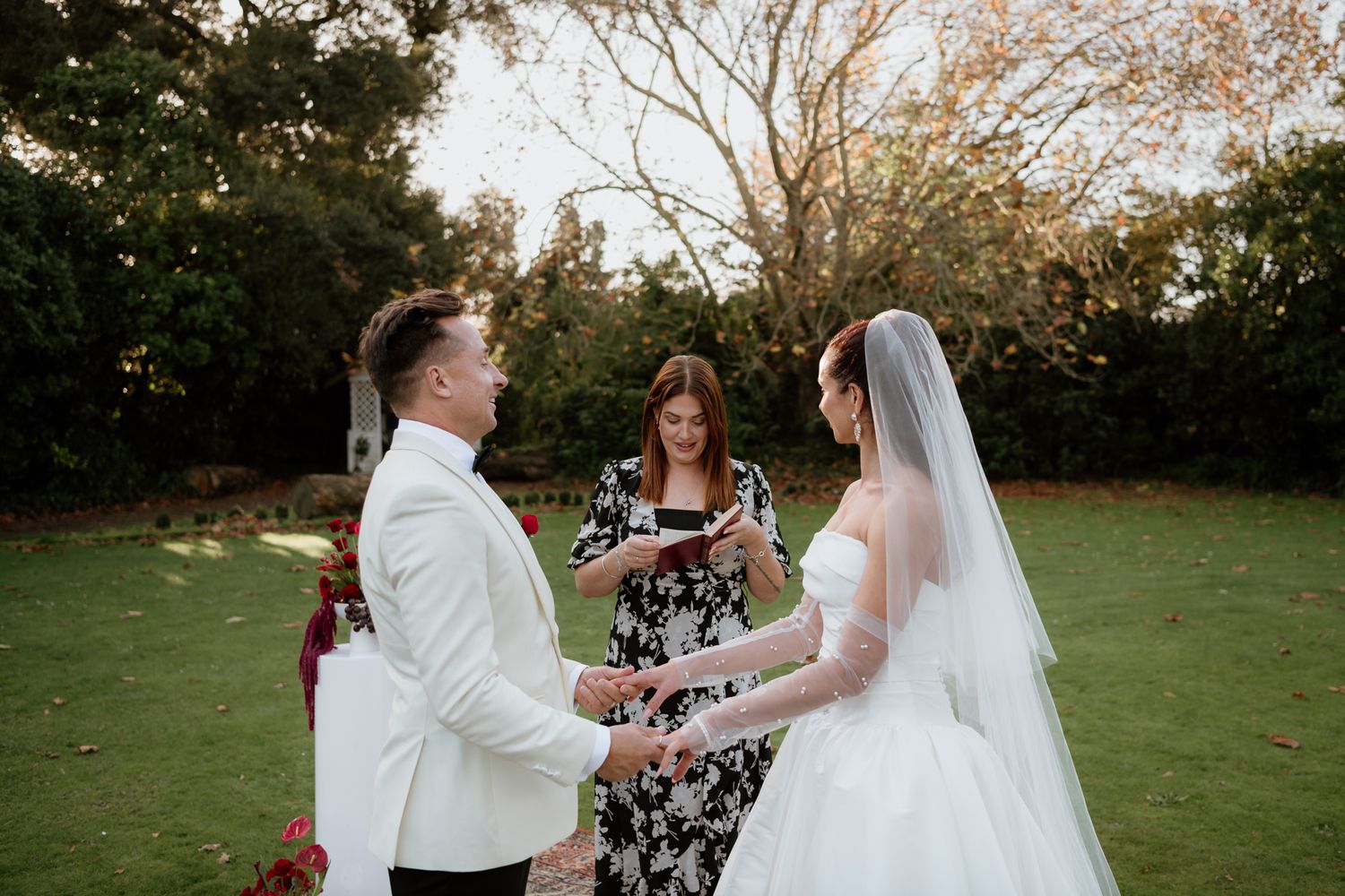 A couple holds hands during an outdoor wedding ceremony with an officiant in a floral dress at Tarureka by August and Vine