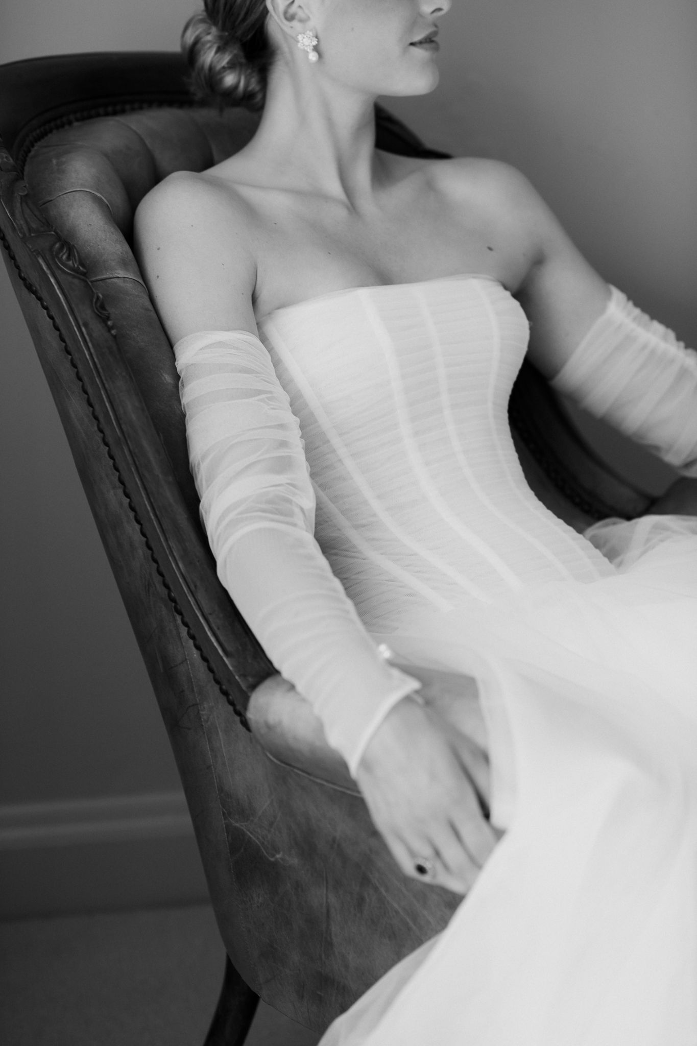 Artistic black and white photo showing bride in corseted elegant white gown reclining against dark antique chair by August and Vine