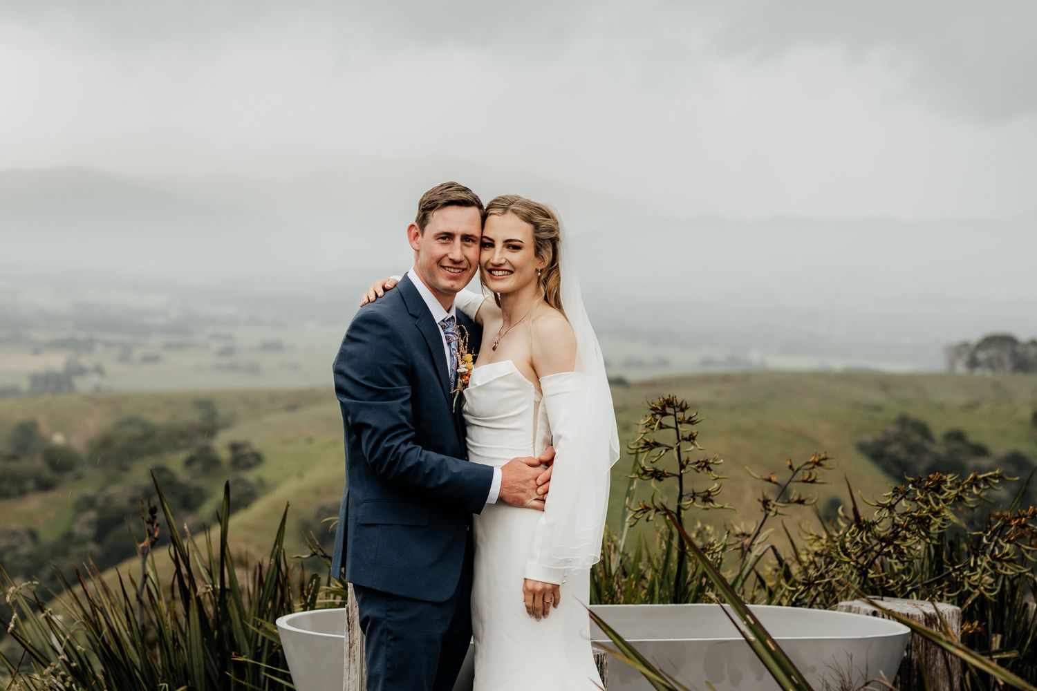 A couple in wedding attire embrace on a scenic hilltop at Whitimanuka overlooking rolling green Wairarapa countryside by August and Vine