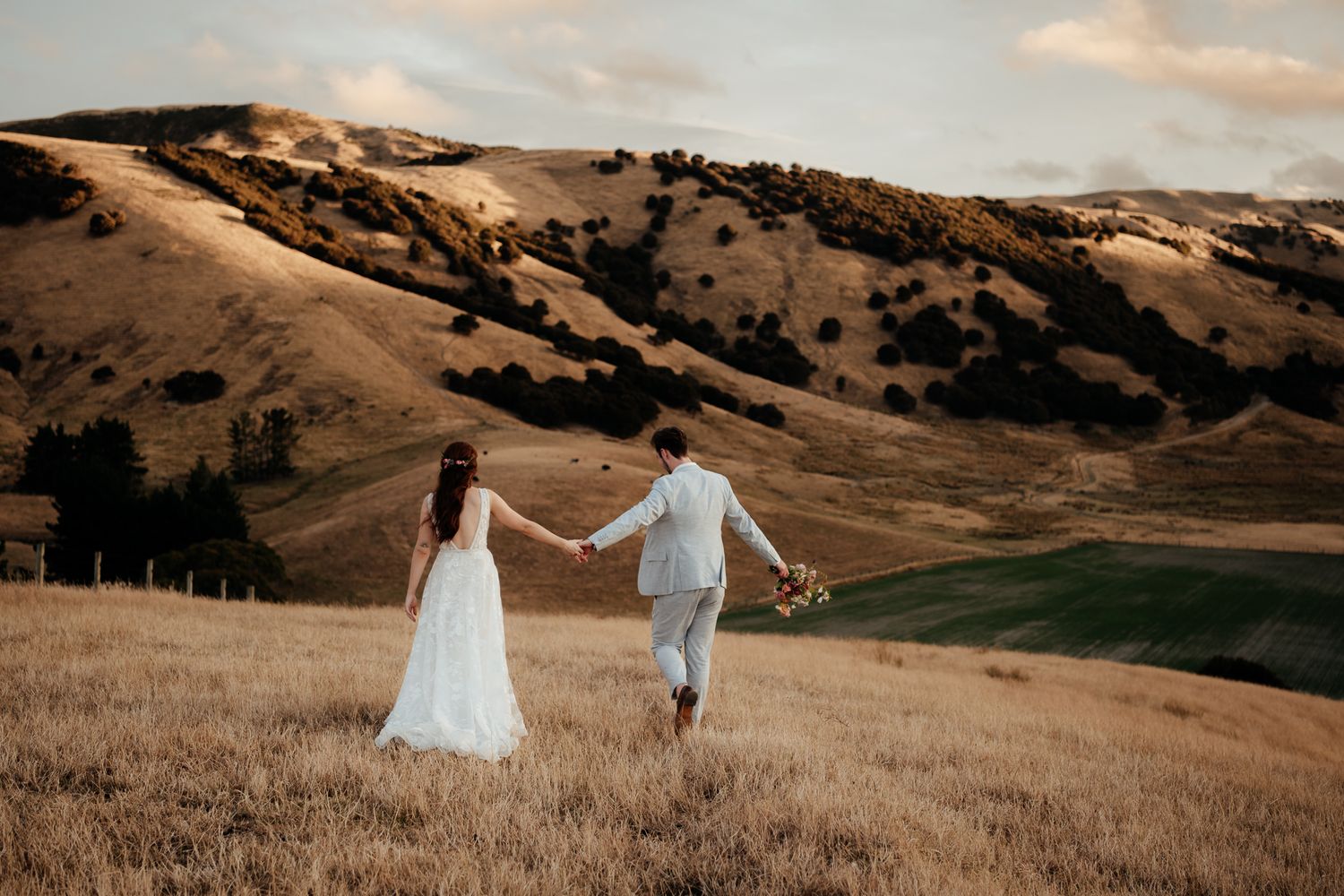 Wedding couple hold hands while walking across golden rolling hills at sunset at Tironui Farm by August and Vine.