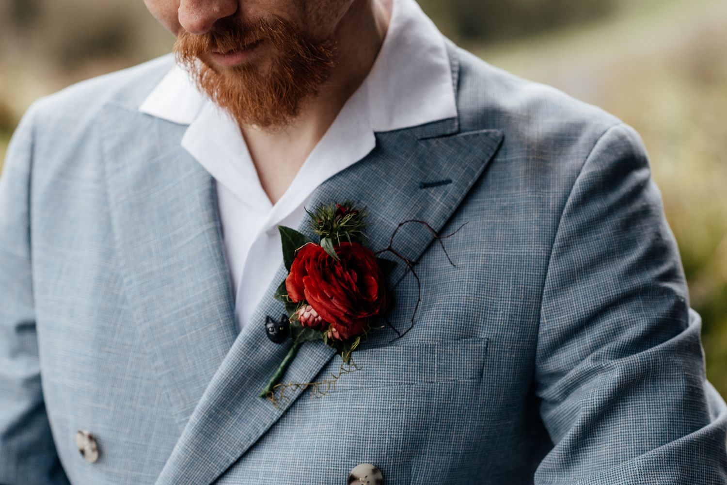 Close-up of a red rose boutonniere pinned to a groom's light blue suit jacket with white shirt by August and Vine