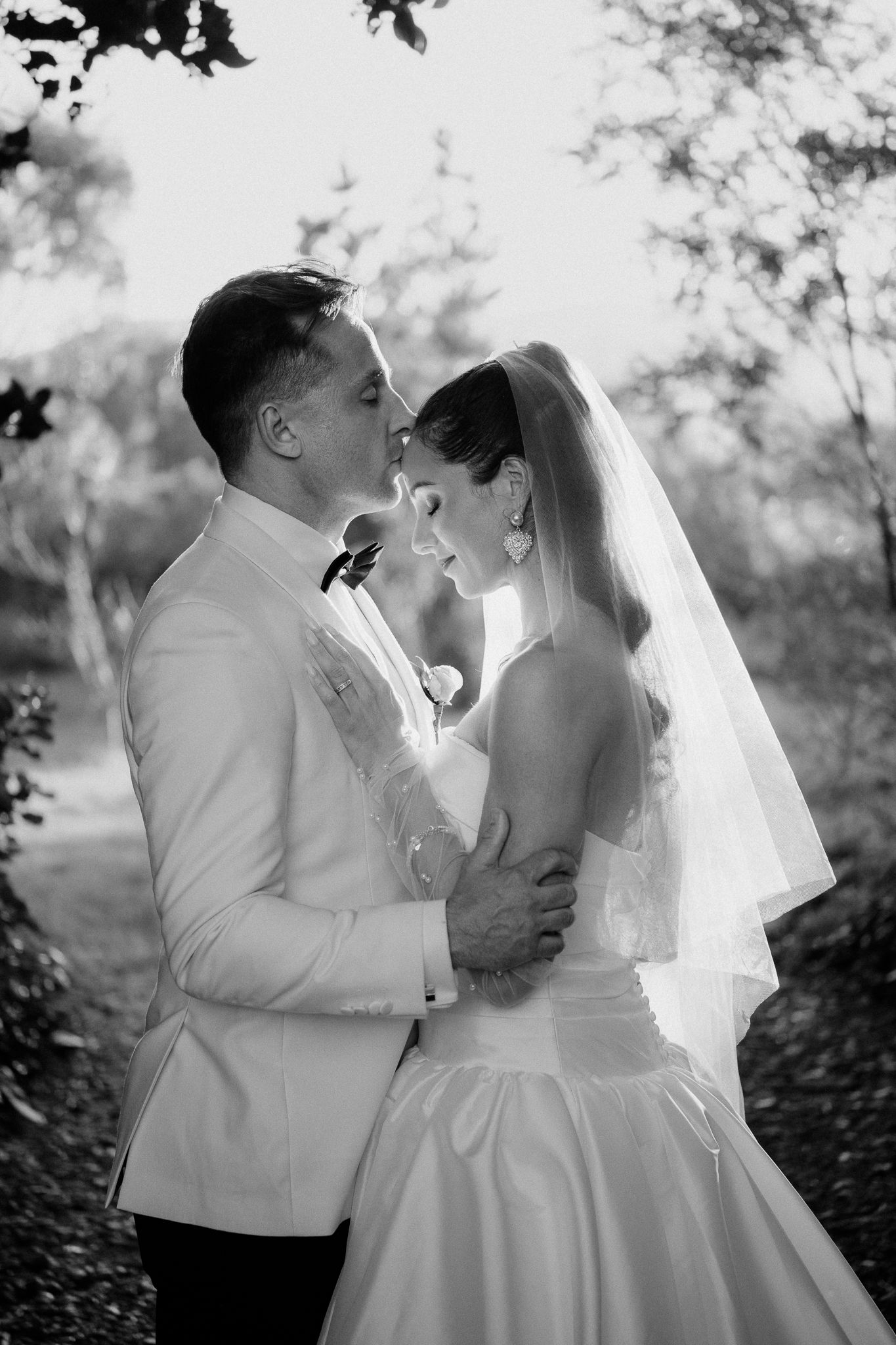 A romantic wedding photo shows a bride and groom in white embracing and kissing in a garden setting by August and Vine