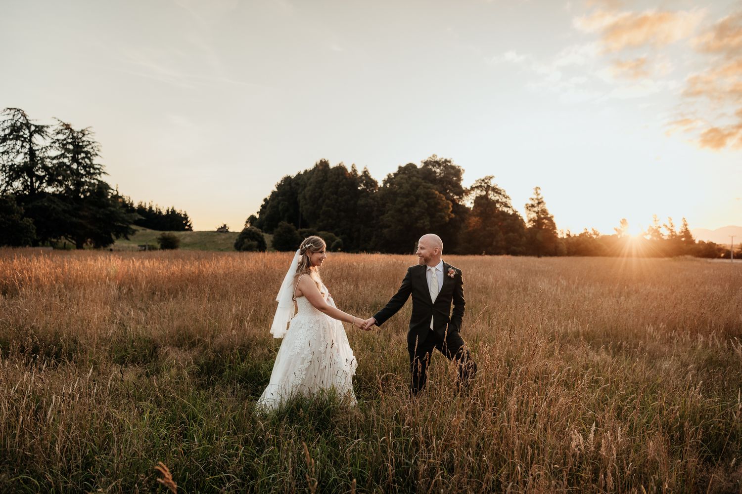 A wedding couple walks through a golden field at sunset holding hands in a romantic outdoor setting at Lacewood Wairarapa by August and Vine