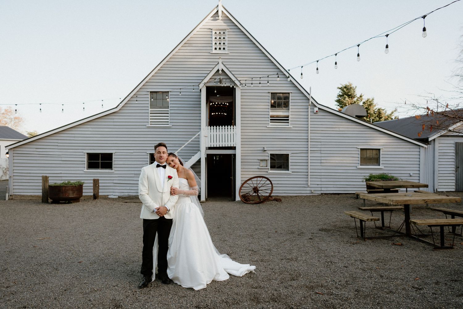 A wedding couple in front of a tall heritage barn decorated with string lights at rustic wedding venue Tarureka at dusk by August and Vine