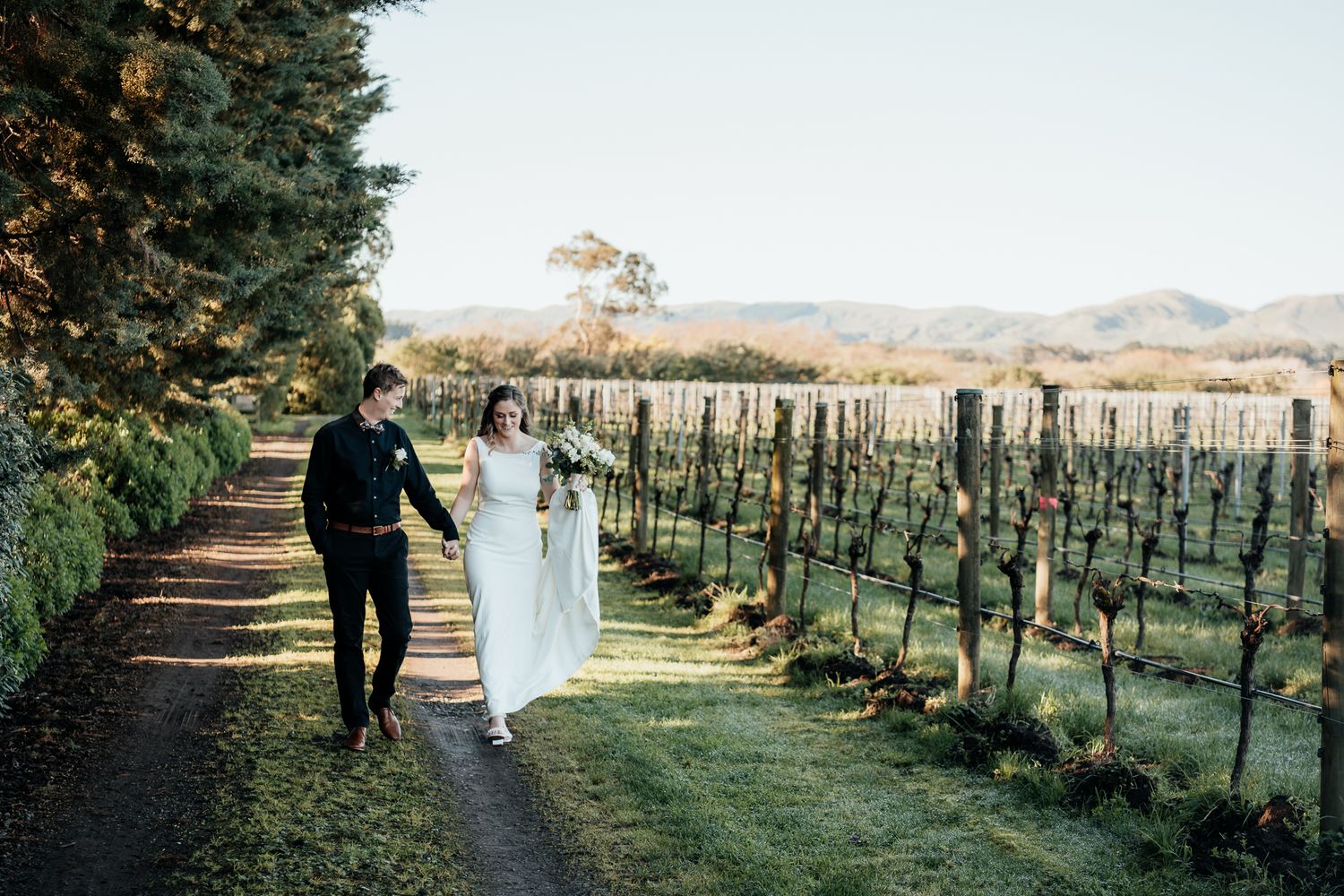 A wedding couple walk hand in hand through a scenic vineyard at Parehua Martinborough on a sunny autumn day by August and Vine