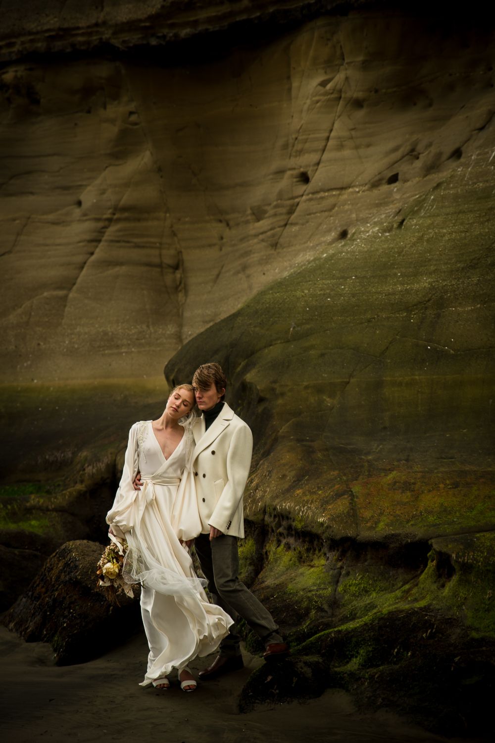 A couple in white wedding attire embrace on mossy rocks against a dramatic cliff backdrop at sunset by August and Vine