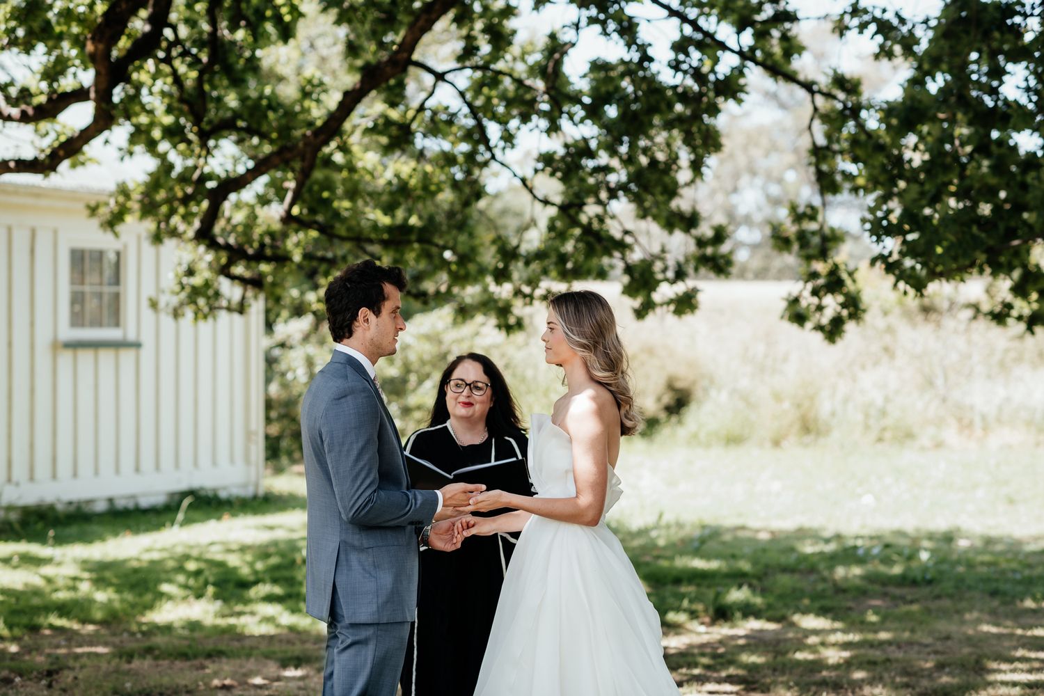 A wedding ceremony takes place outdoors under a large tree by August and Vine