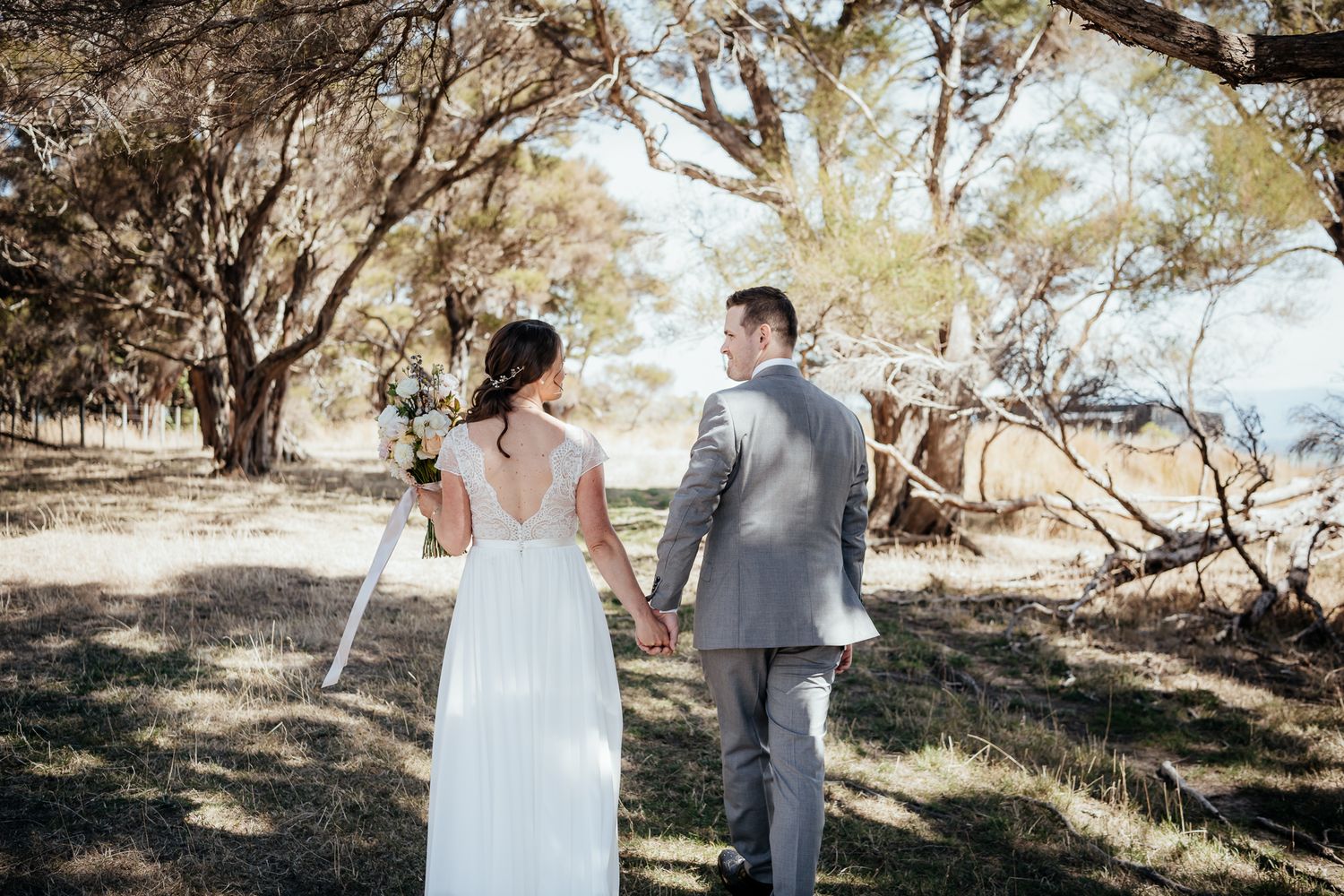 Couple holds hands while walking through sun-dappled Manuka in elegant wedding attire at Whitimanuka by August and Vine