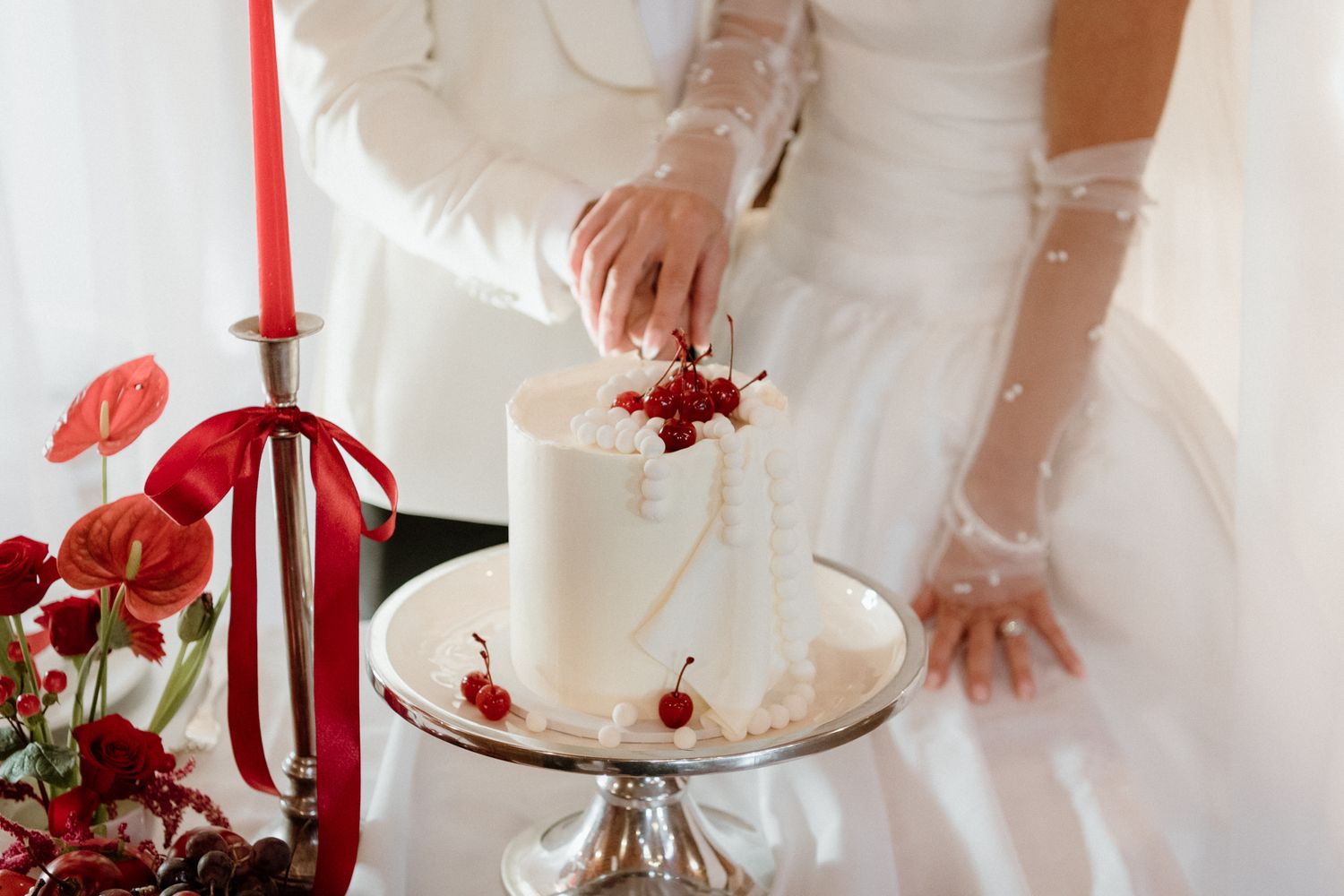A couple cut their modern small wedding cake on a table set with red candles, red flowers and white linens by August and Vine