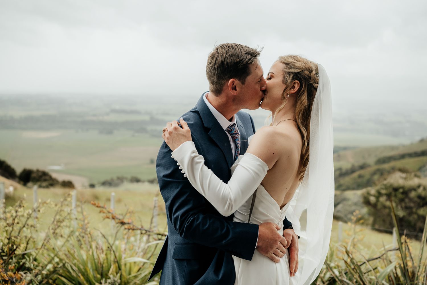 A romantic wedding couple embrace in front of rolling green hills and scenic Wairarapa countryside views by August and Vine