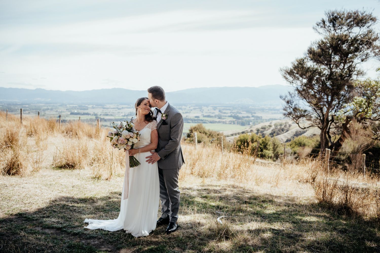 A scenic wedding moment as couple embraces during outdoor ceremony with rolling hills in the distance at Whitimanuka by August and Vine