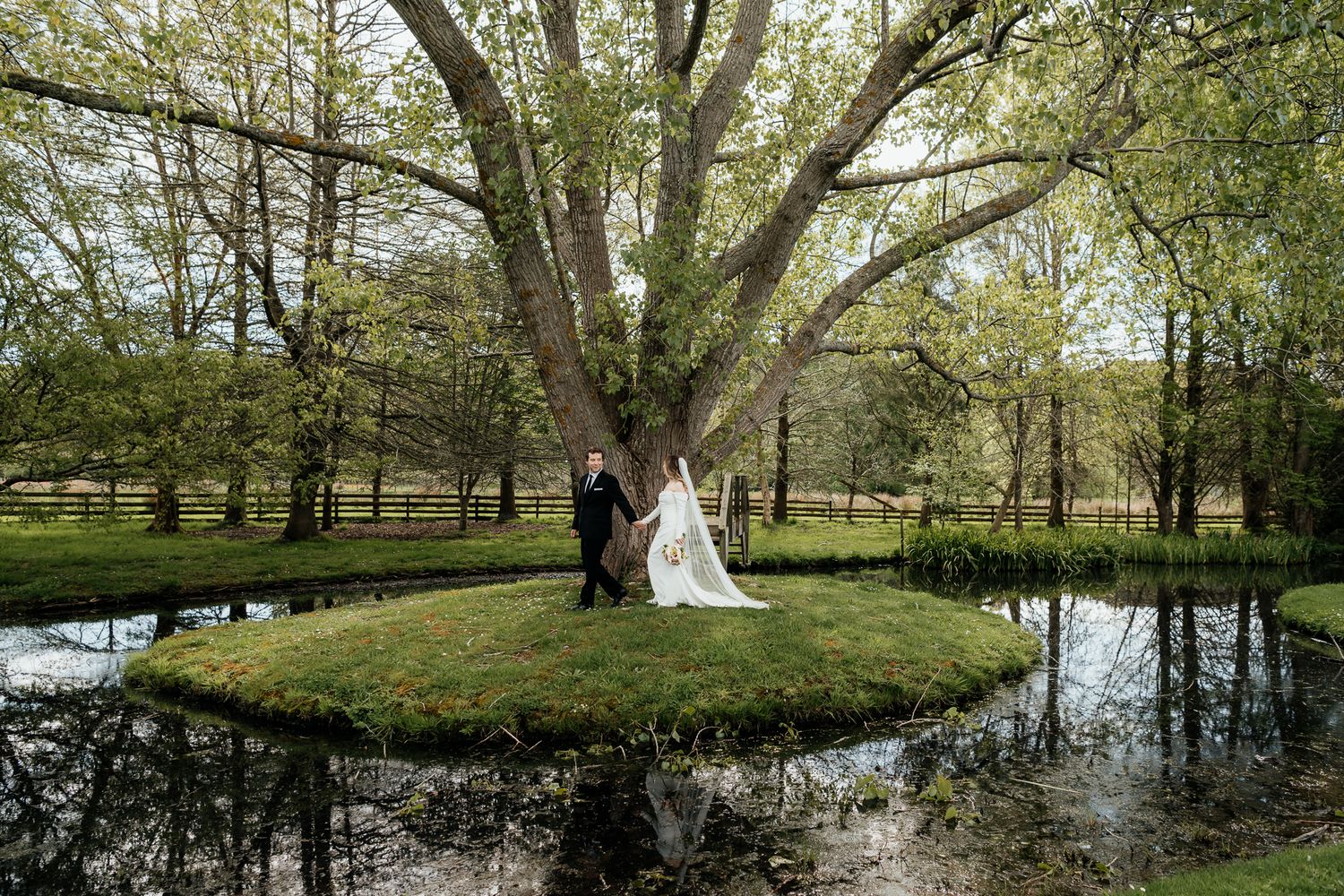 A wedding couple stand beneath a large tree on a small island surrounded by reflective water by August and Vine