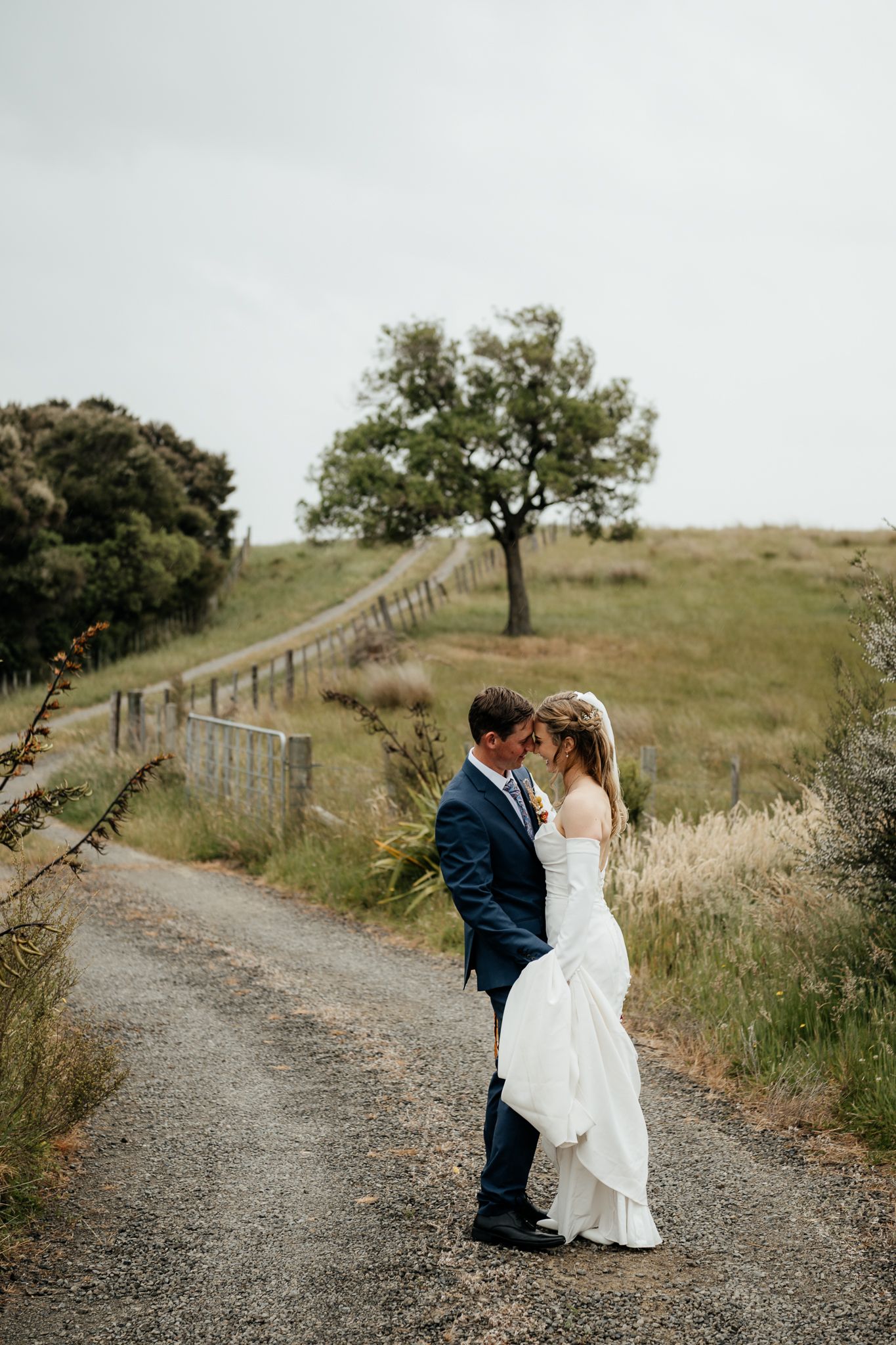 Romantic couple embrace on a winding gravel path surrounded by rolling Wairarapa hills and rustic wooden fencing by August and Vine