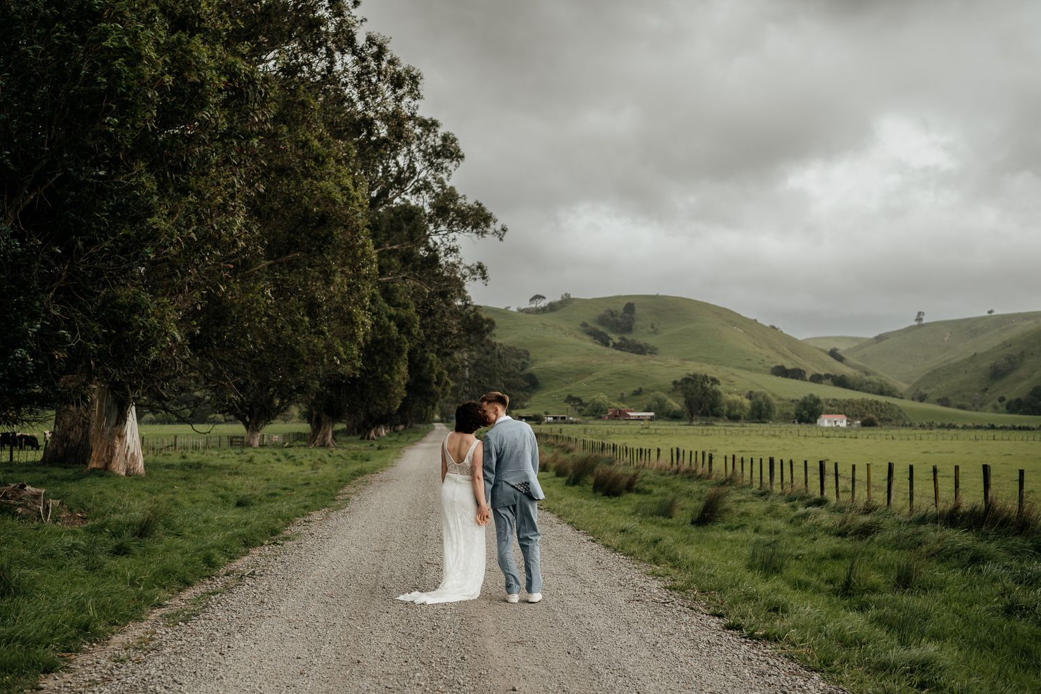 A couple walks along a scenic rural gravel path surrounded by rolling green hills and pastoral farmland by August and Vine