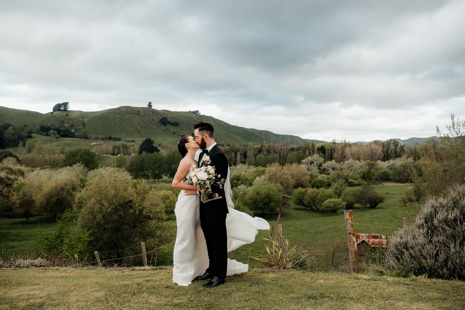A wedding couple embrace on a hilltop overlooking a scenic country landscape with dramatic cloudy skies at The Runholder by August and Vine