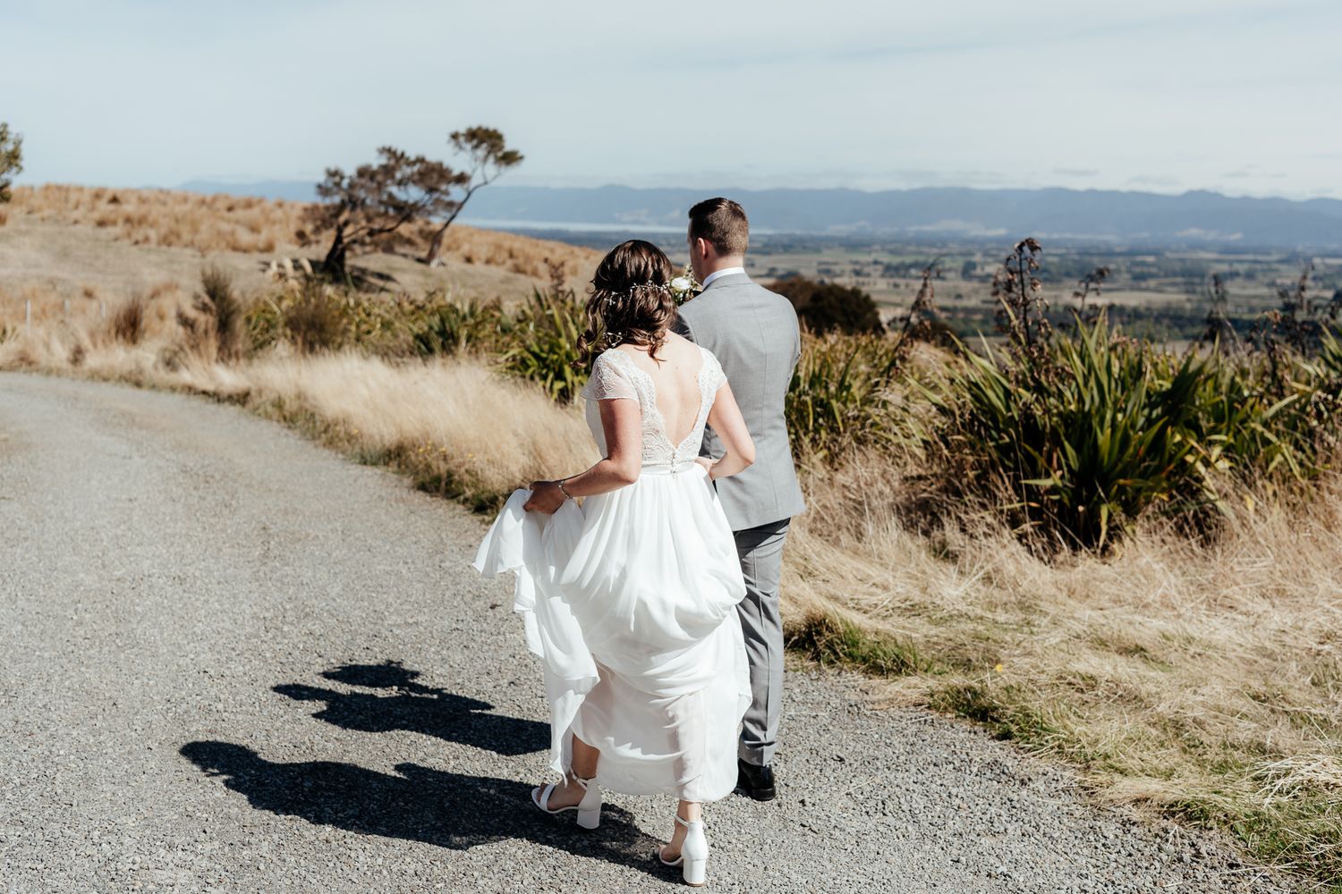 A couple in wedding attire walks along a scenic ridgeline with mountains in the distance at Whitimanuka by August and Vine