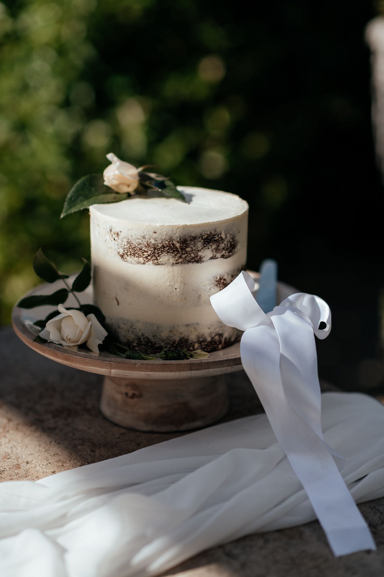 A small semi-naked wedding cake decorated with greenery and white ribbon on a wooden cake stand by August and Vine