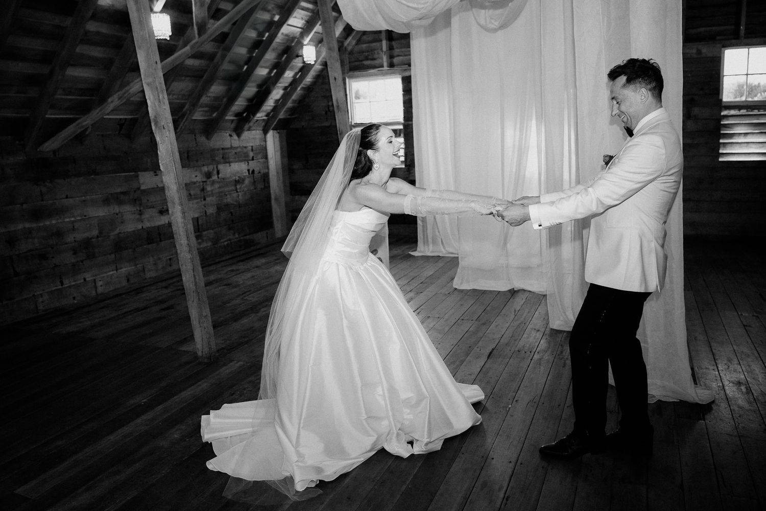 A couple in wedding attire dance together in a rustic barn, creating a romantic black and white moment captured by August and Vine