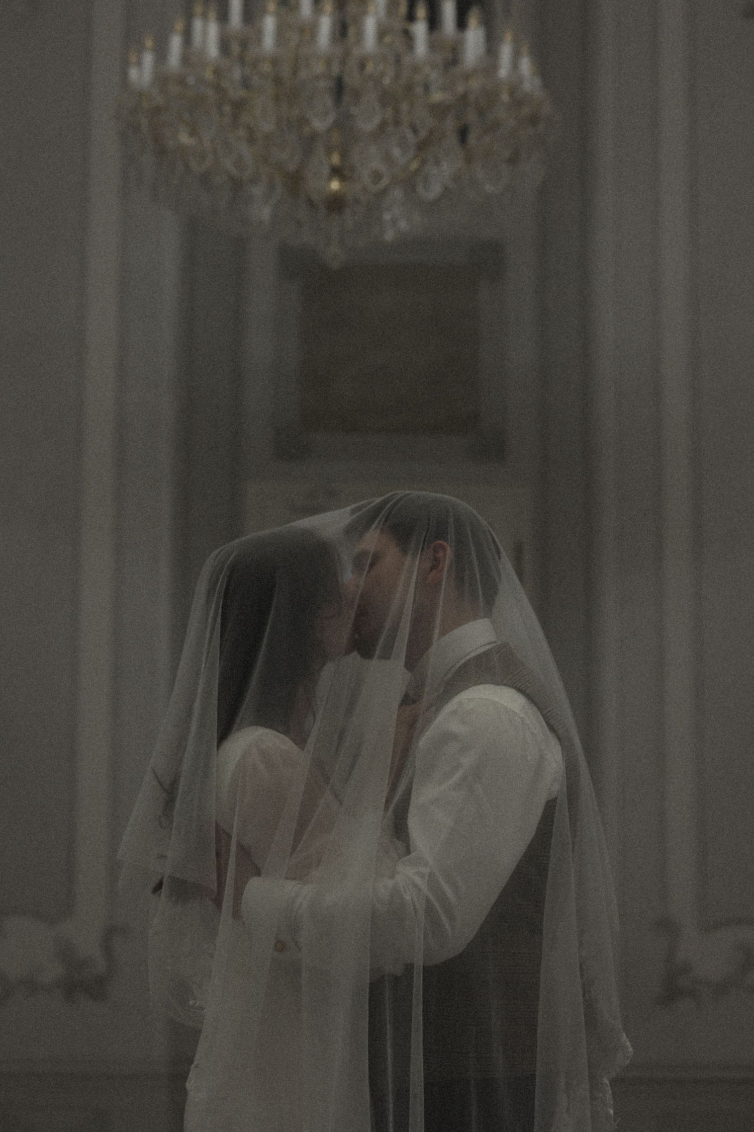 A romantic wedding couple shares a tender kiss under a flowing white veil in an elegant room with chandeliers.