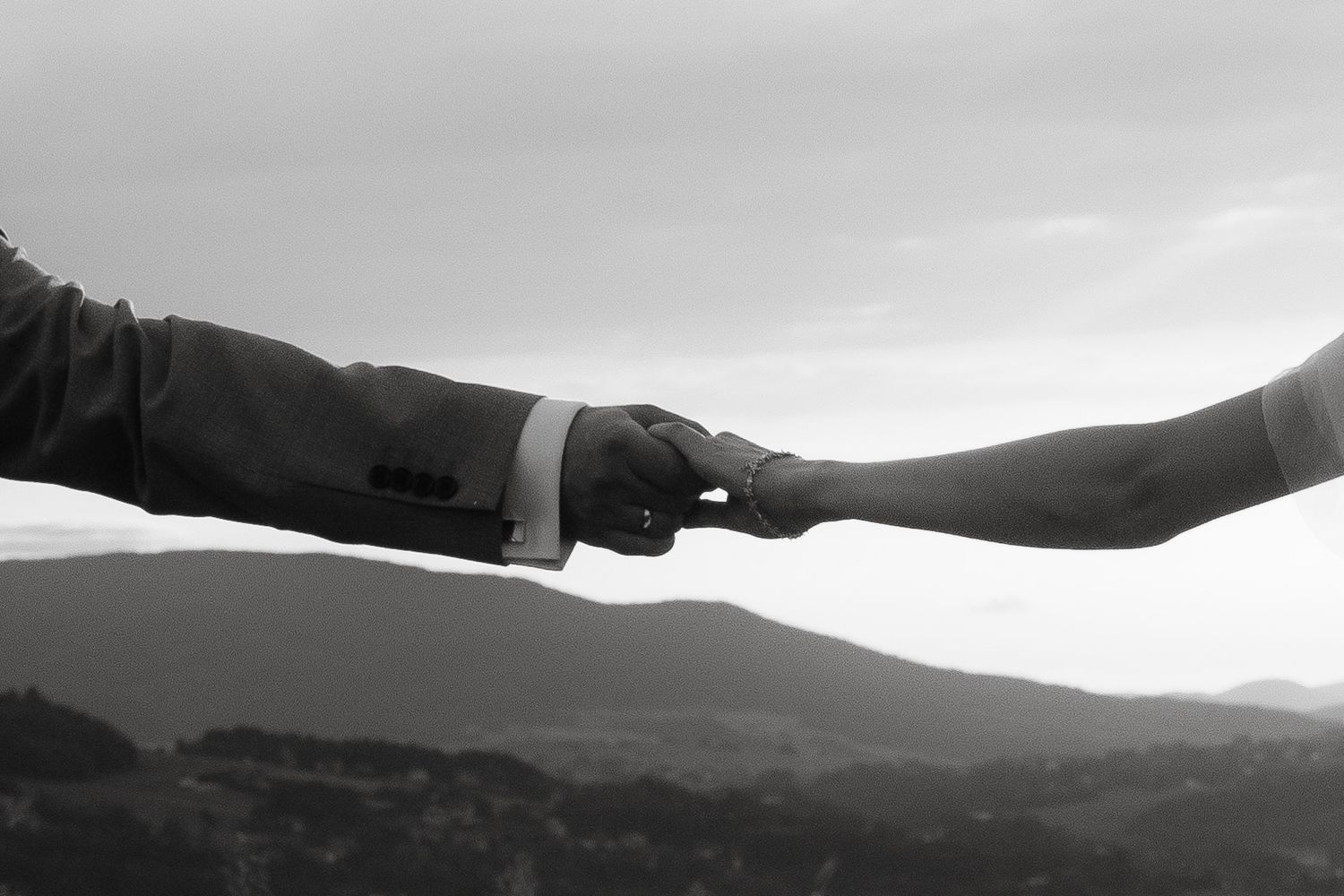 Black and white photo of two hands reaching out to hold each other against a blurred mountain landscape background.
