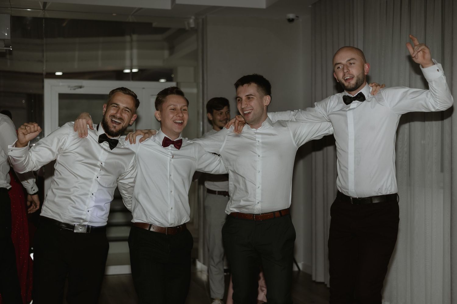 Groomsmen in white shirts and bowties celebrating joyfully at a wedding reception.