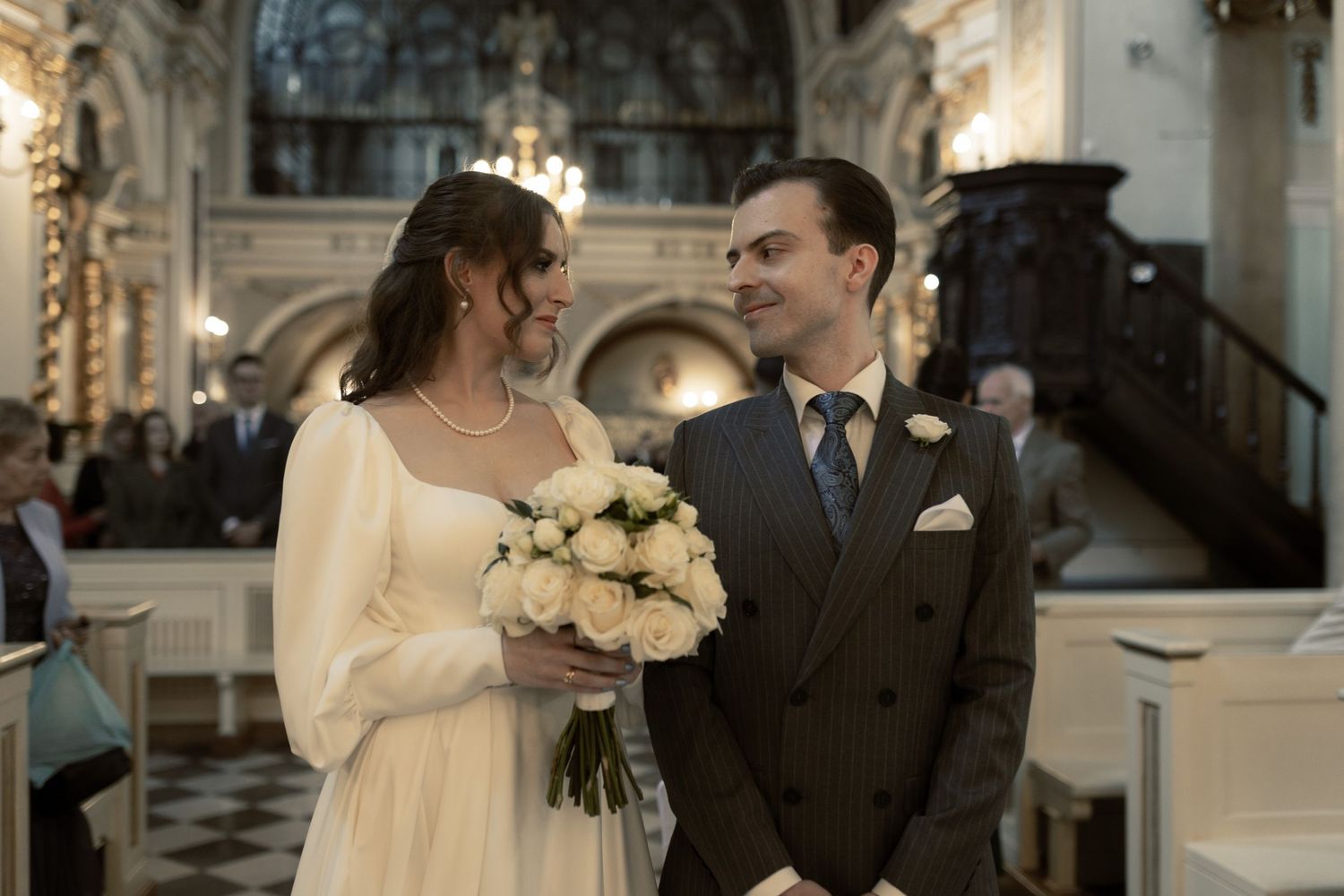 A couple stands together during their wedding ceremony in an ornate church interior while holding white roses.