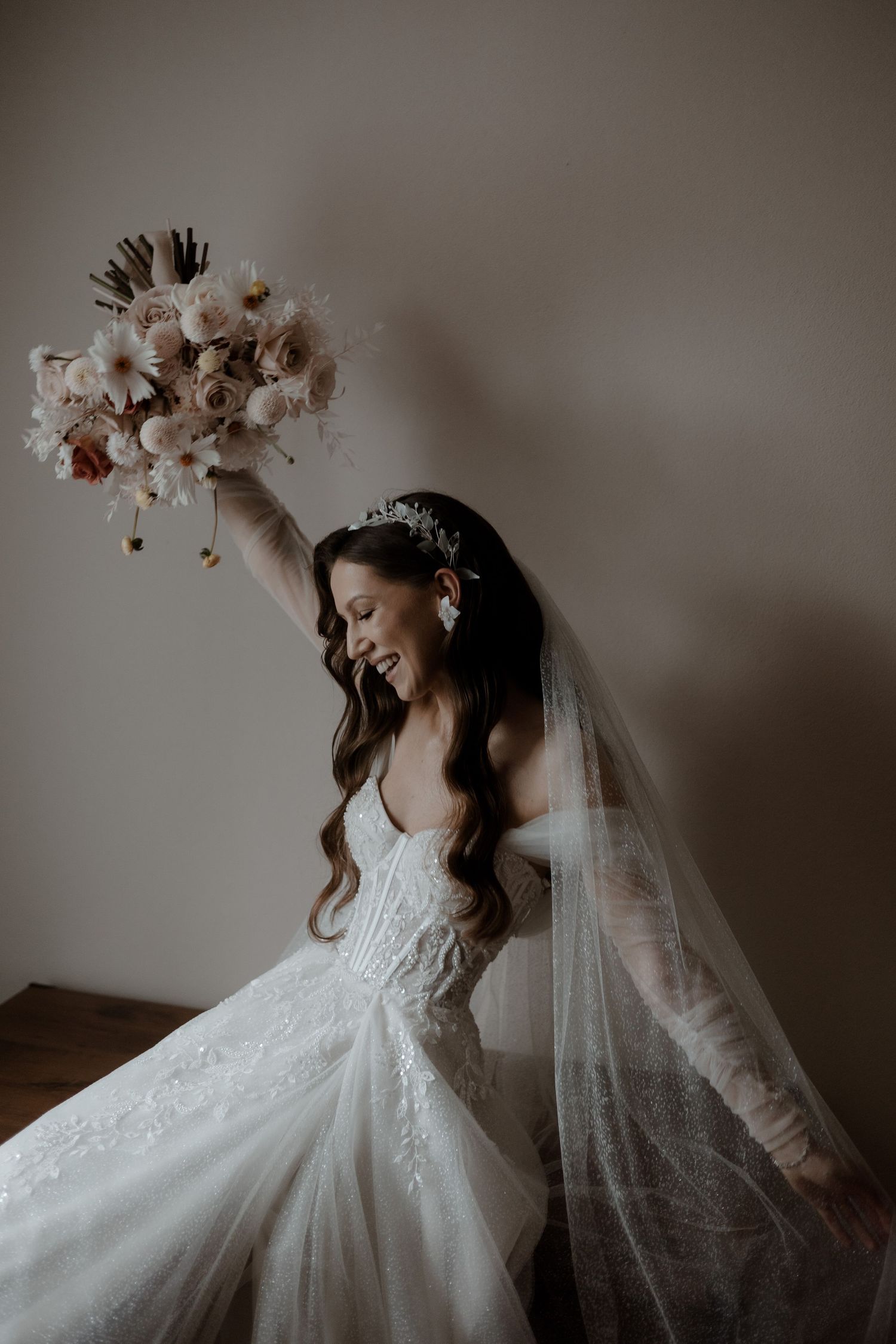 Elegant bride in white lace gown and flowing veil holds up a romantic bouquet of dried flowers while posing.