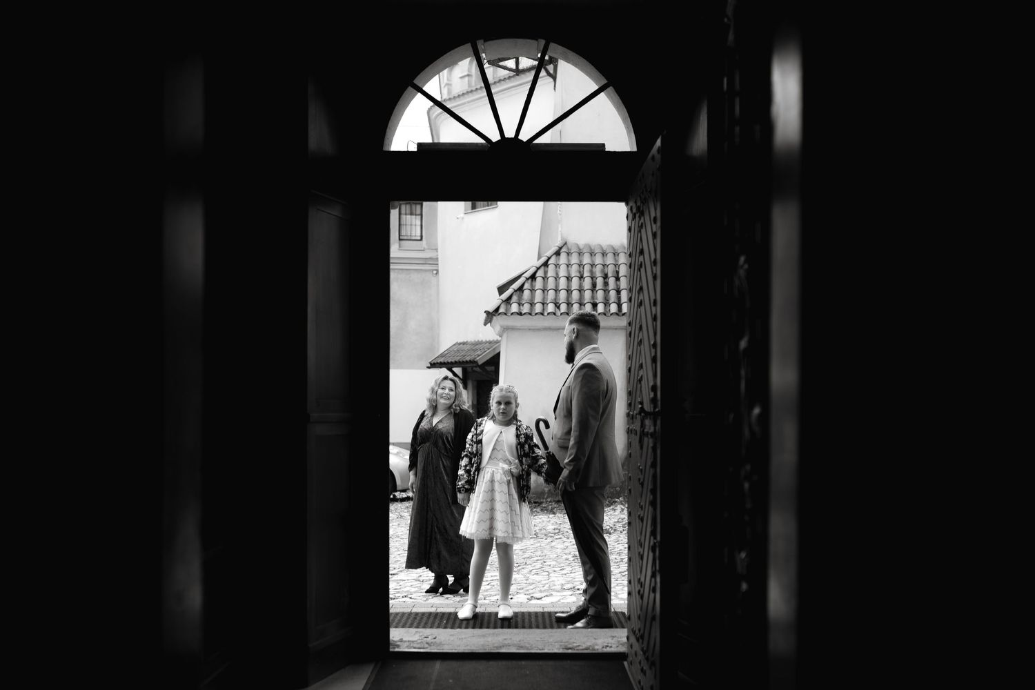Through a dark doorway with an arched window, three silhouettes of people conversing on a porch are seen in black and white.