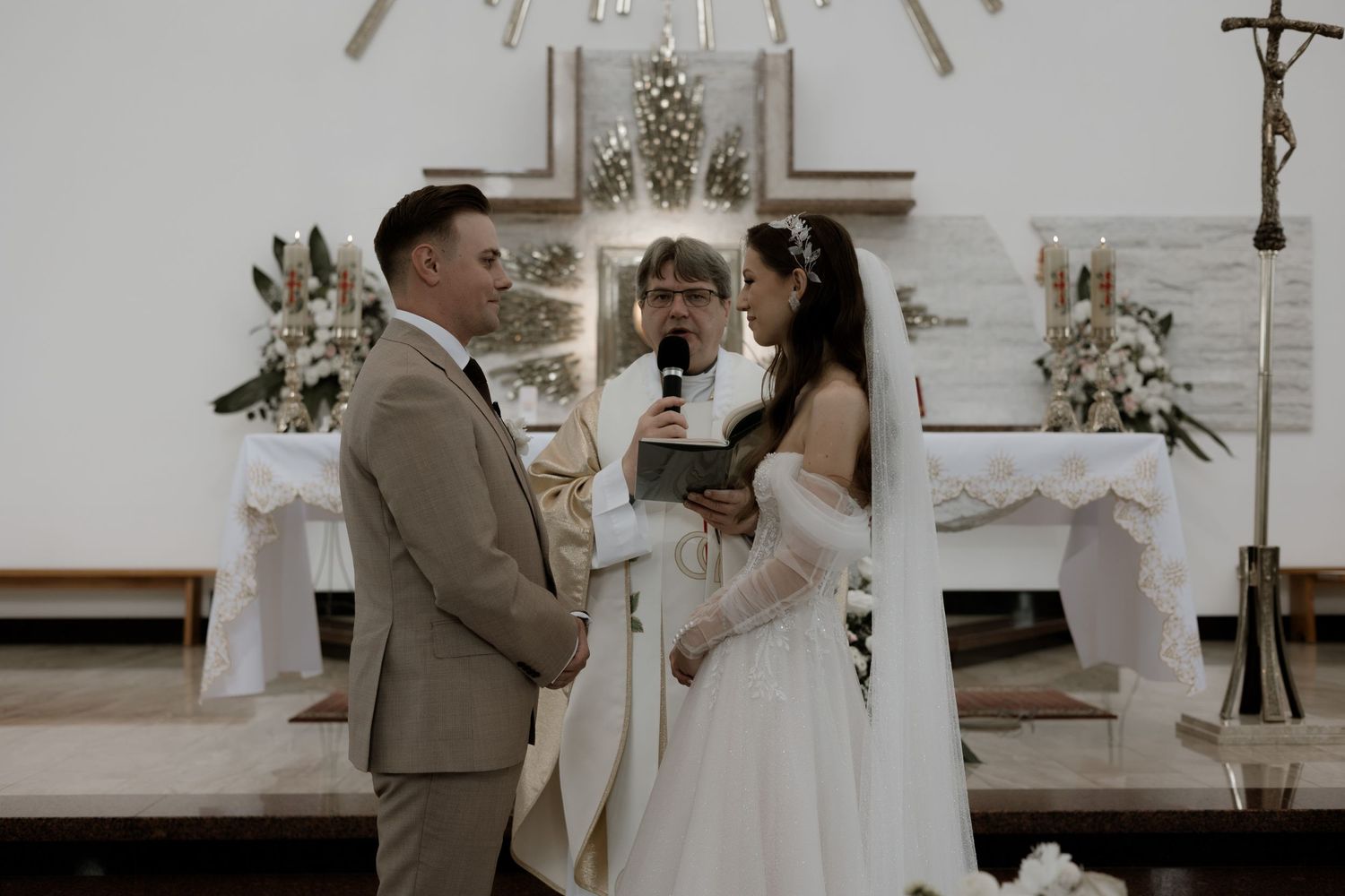 Wedding ceremony takes place in a church with white floral arrangements and decorative details.