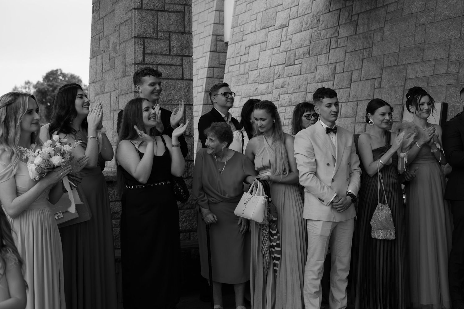 A black and white photo of a group of formally dressed people standing in a line next to a stone wall.