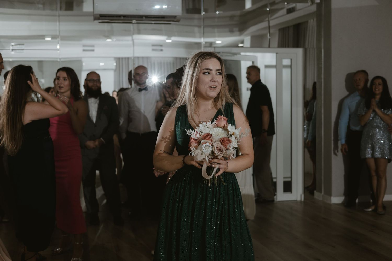 A person wearing glasses and a green dress holds a bouquet at an indoor event while others mingle in the background.