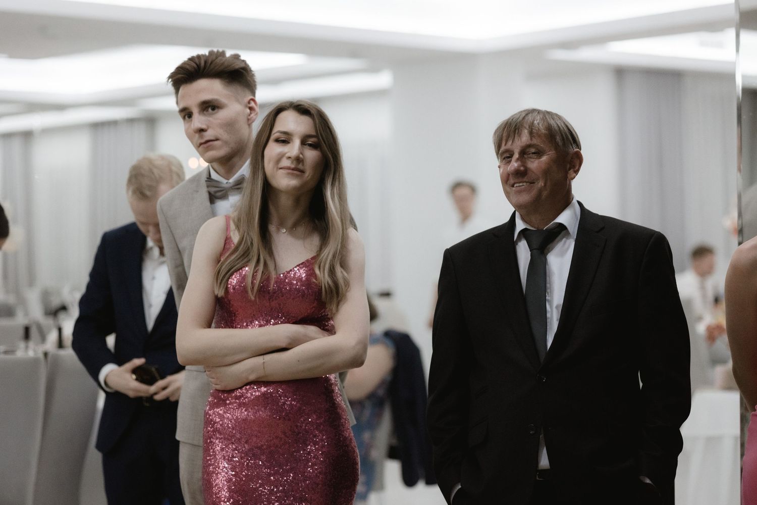 Three people stand in a formal setting with one wearing a sparkly red dress and others in suits at an event.