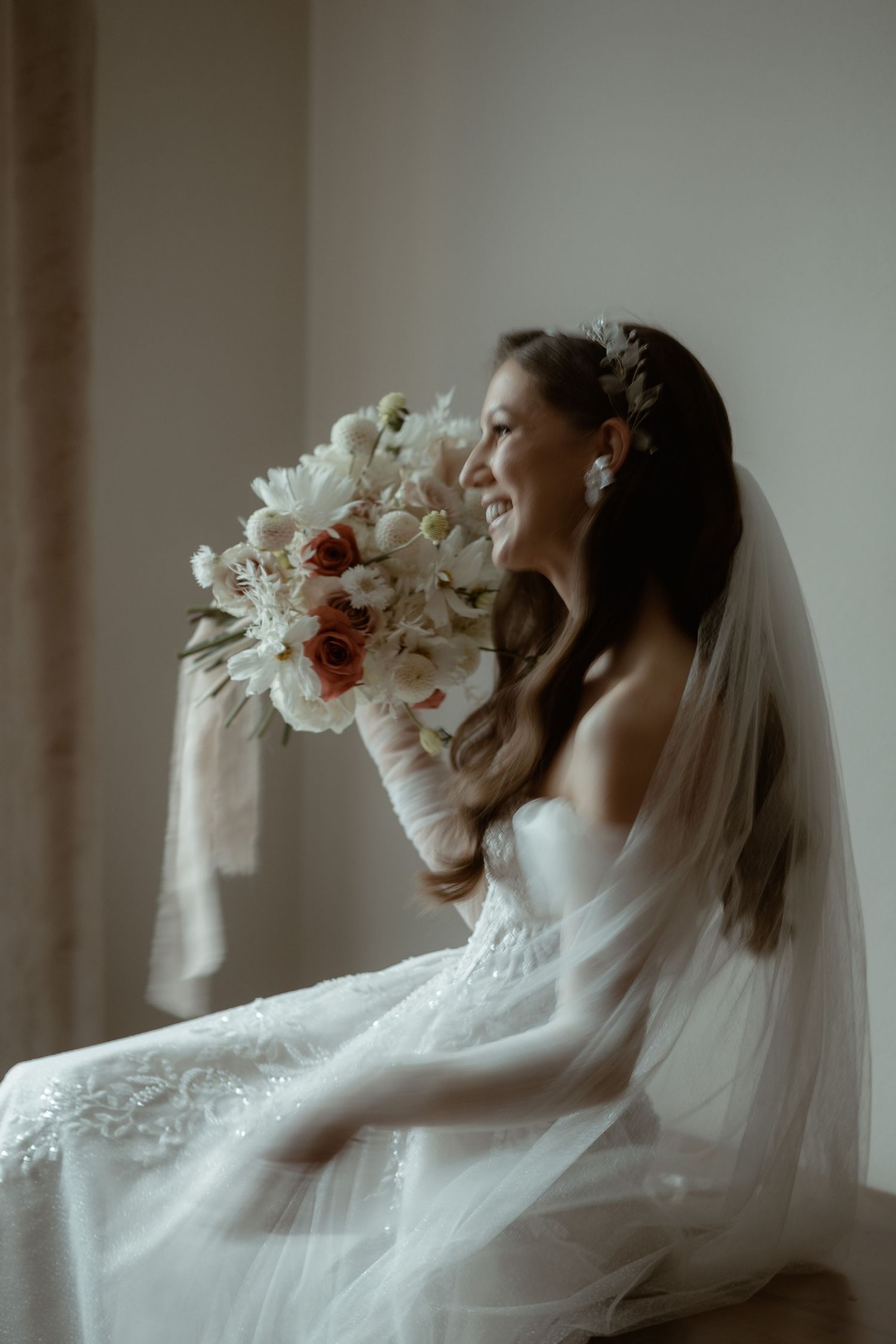 A bride in a white gown and veil holds a beautiful bouquet of white and red flowers while sitting in soft window light.
