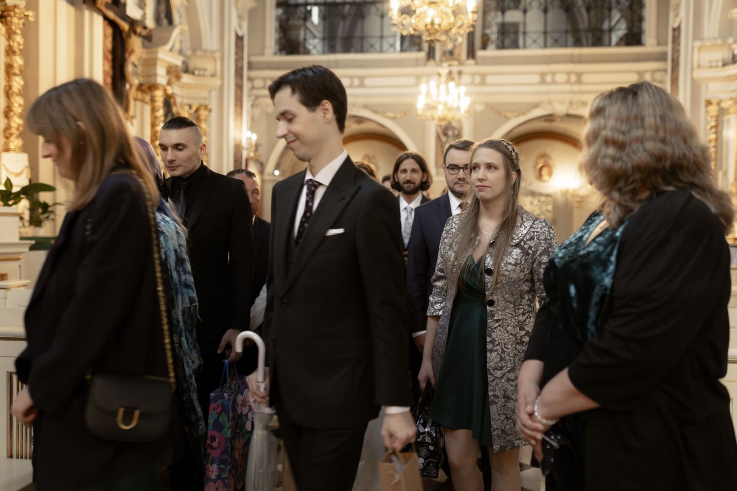 A group of formally dressed people gather in an ornate room with chandeliers and elegant architecture.