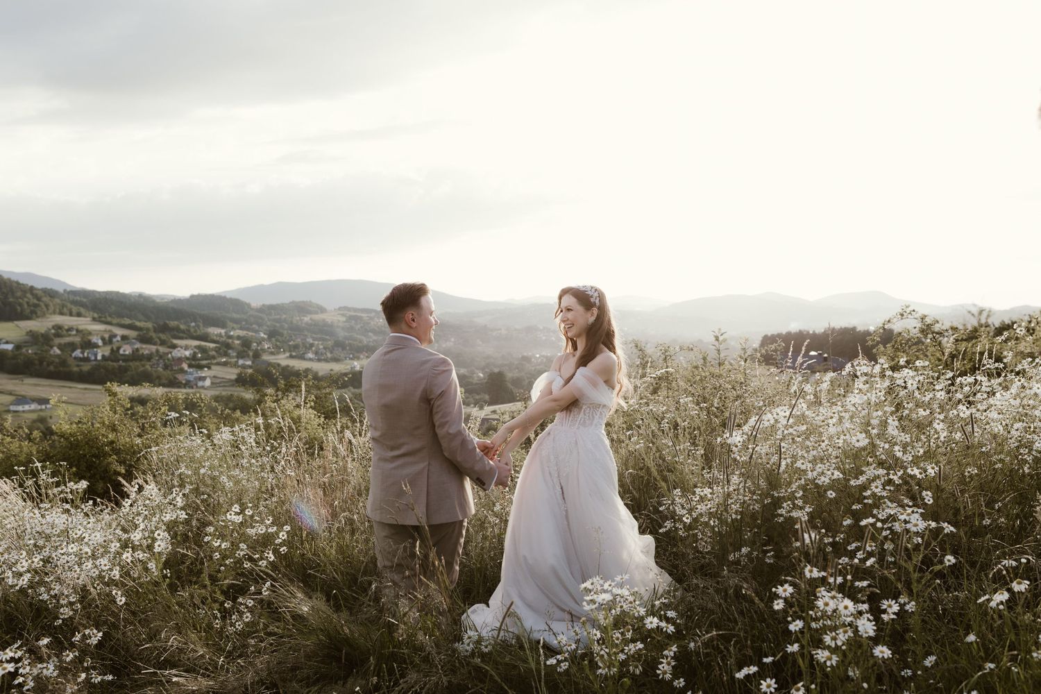 A backlit couple stands together in a meadow with wildflowers during sunset with mountains in the background.