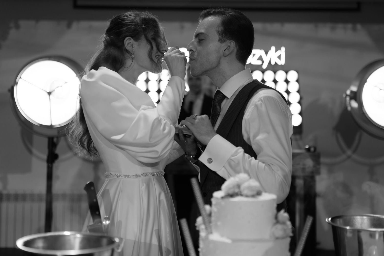 A romantic moment between a couple in a black and white photo as they feed each other wedding cake in front of light fixtures.