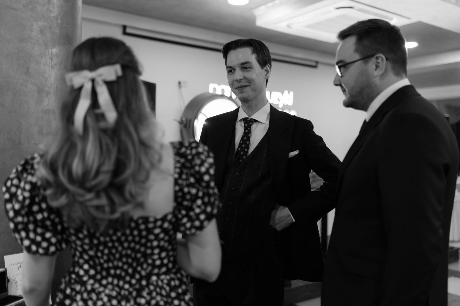A black and white candid shot of people chatting at an indoor gathering, with a person in polka dots facing away from camera.