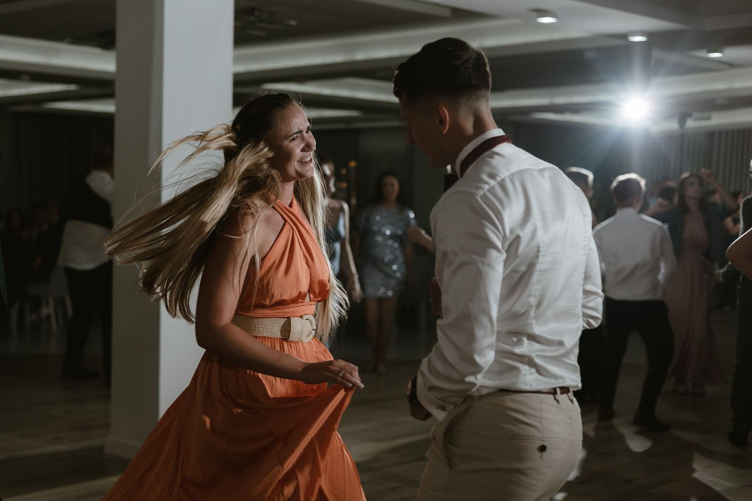 Two people dancing together at a wedding reception, with a woman in a flowing coral dress and a man in a white shirt.